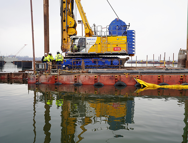 men stand next to a drill on a platform in a river on a cloudy day